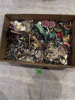 Top-down view of cardboard box filled with assorted costume jewelry items including necklaces, bracelets, earrings, brooches, and loose beads of various colors and materials.
