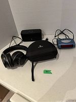 Three electronic items on white table: black Vankyo Bluetooth headphones with carrying case, black Doss Bluetooth speaker, blue Cosmo alarm clock.