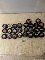 Photo showing 26 hockey pucks arranged in rows on a surface, displaying a variety of team logos and brands.