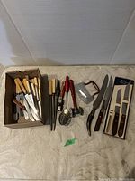 Photo displaying various knives with cream handles, red handled kitchen tools, metal scraper, and boxed kitchen knives on a beige cloth background