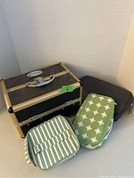 One photo showing a hard shell makeup case in black and gold finish alongside three soft makeup bags in green and white stripes, green and white circle pattern, and solid black.