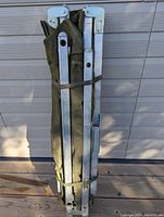 Folded aluminum military cot leaning against a garage door; showing metal frame and green canvas fabric secured with straps.