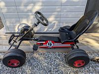 Side angle view of black pedal car showing plastic seat, steering wheel, pedals, and four wide tires with red rims.