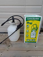 White plastic lawn and garden sprayer with pump handle, black hose, and sprayer wand next to original box.
