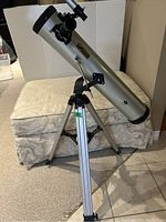Full view of silver Optics brand telescope mounted on tripod with legs extended indoors on carpet.