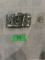 Front view of the metal belt buckle showing three detailed Tiki god faces with different emotions and expressions. Coin for size reference.