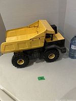 Side view of the yellow and black Tonka metal dump truck next to a water bottle for scale showing the condition and details of the truck body and wheels.