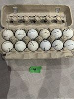 Set of 12 mixed used golf balls displayed in an open cardboard egg carton-style holder. Balls show varied brand names and scuff marks from use.
