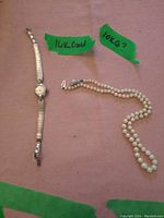 Watch and pearl necklace displayed on pink background with green tape indicating gold markings
