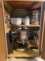 Cookware items inside a wooden cabinet, including white ceramic bowls on upper shelf, a plastic container, and stainless steel pots and pans on the lower shelf.