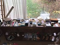 Wide view of assortment including watches, earrings, decorative box, perfume bottle, and other costume jewelry on a wooden table by window.