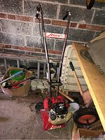 Full view of red Honda soil tiller with metal handles, tines, and fuel tank in garage environment.