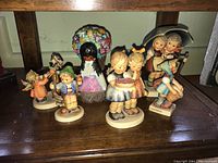 Photo of six figurines, five Hummel figurines depicting children and one Degrazia Flower Girl figurine holding a colorful flower bouquet.