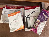 Photo of four OttLite lamps in their original boxes for sale in the lot: two Reader's HD rechargeable portable book lamps, one Creative HD craft lamp, and a universal craft lamp with clip.