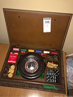 Photo of the open wooden box showing roulette wheel at center with chips, cards, chess pieces, domino tiles, and dice around.