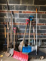 Full view of all garden tools including shovels, rakes, brooms, pitchfork, and snow shovels leaning against garage wall