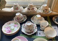 Overview of ten teacups and saucers, sugar bowl and creamer placed on a window ledge showing various floral patterns and gold trim