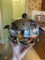 Front view of the West Bend Penguin stainless steel serving server with black knob lid, showing shiny reflective surface and side handles held in hand