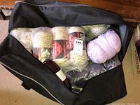 Open black hockey bag revealing multiple skeins of yarn, some wrapped in clear plastic, with different colors including purple and flecked varieties.