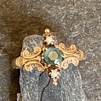 Overhead view of ring lying flat, showing central green faceted stone, flanking seed pearls, and scrolled frame.