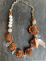 Full view of asymmetrical necklace showing amber crystal clusters, white beads, crystal spheres, shell pieces, and gold-tone chain.