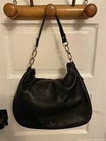 Black slightly textured leather shoulder bag hanging on wooden pegs, showing overall front shape with chain and leather strap handle.