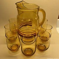 Amber glass pitcher with rust and amber horizontal stripes around the middle, surrounded by seven matching amber glass tumblers.
