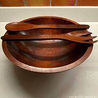 Maple wood bowl with wooden spoon and fork utensils arranged inside, front angle view showing bowl's curved shape and wood grain.