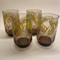 Four smoky brown glasses decorated with golden wheat sheafs and tangerine leaves shown from the front on a white background.
