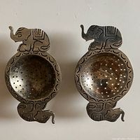 Top down view of the pair of silver plate tea strainers with elephant handles showing the perforated tea bowls and elephant motifs on ends.