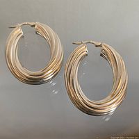 Two sterling silver chunky twisted oval hoop earrings shown front side on a flat surface with reflective background, showing shape and design clearly.