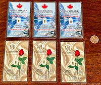 View of all six gold bullion bars arranged on a wooden surface, three with a blue and white maple leaf design and three with a satin gold and red rose design, with a penny for scale