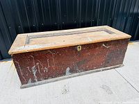 Large antique wooden carpenters tool chest shown from front and side angles on concrete with a dark background. Chest has worn wood finish and paint splatters. Metal carry handles on sides and a lock hole on front.