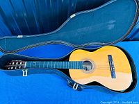 Open classical guitar inside black hard case on blue surface, showing guitar full body and foam interior of case.