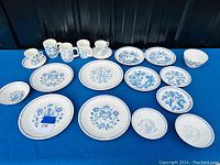 Overview photo showing the full set of ceramic cups, saucers, mugs, and plates arranged on a blue surface.