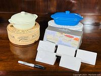 Two stoneware mini casseroles, one cream/off-white and one bright blue, displayed on a wooden surface with their original boxes. Six white rectangular place setting cards stand in front, accompanied by a black marker for writing on the cards.