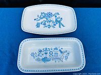 Two similar rectangular ceramic platters with rounded corners on blue background, showing the blue Lotte folk art pattern with figures and flowers