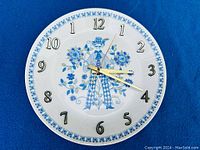 Full view of the Figgjo Flint Turi Design Lotte plate clock on a blue background. Clock face shows the central blue figure with flowers, black numerals, and gold-tone hands.