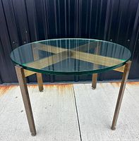 Front angle view of the round glass top coffee table showing the bronze-coated metal legs and base structure on concrete ground.