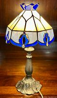 Frontal view of the Tiffany style stained glass desk lamp showing the bronzed ornamental base and the lit stained glass shade featuring white and blue glass panels with floral motifs.