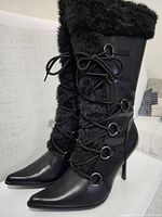 Side angle of left boot showing black faux fur front panel with lace-up detail through silver metal rings, pointed toe, and stiletto heel.