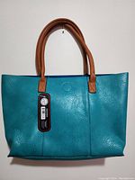 Front view of teal leather handbag with brown leather handles hanging on wall, showing texture and stitching details.