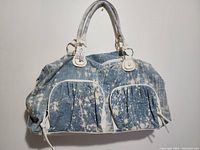 Front view of distressed denim hand bag with white splatter and two zipped front pockets with white trim and leather-like handles.