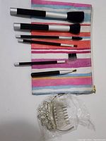 Six makeup brushes with black and silver handles displayed on colorful striped zippered bag, plus clear plastic hair comb clip with rhinestone decoration wrapped in plastic.