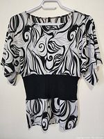 Front view of the black and white decorative women's top on a hanger showing the swirl pattern and fitted black waistband.