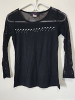 Front view of the black women's top on hanger showing mesh fabric on sleeves, upper chest and decorative silver studs.