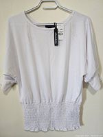 Full view of the white women's top, showing boat neckline, 3/4 sleeves, and hanging on a hanger with tag attached.