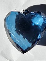 Close-up photo showing the front side of the teal-colored faceted heart pendant with reflective surface and metal loop attachment.
