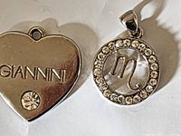 Two silver-tone pendants shown together, one circular Scorpio zodiac style with rhinestones, one heart-shaped with GIANNI engraving and rhinestone.