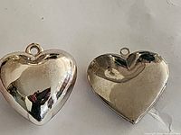 Photo shows two heart-shaped chrome pendants, one smooth and polished, the other with embossed floral decoration and smaller heart in center.